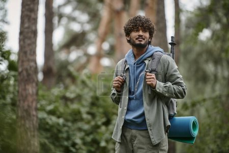 Photo for Pleased young indian traveler holding backpack and looking away in forest - Royalty Free Image