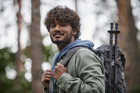 portrait of smiling indian hiker holding backpack and looking at camera in blurred forest