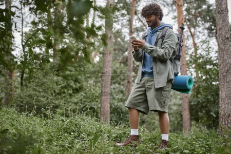 sonriente joven turista indio con mochila usando teléfono inteligente mientras está de pie en el bosque