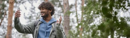turista indio positivo con mochila que tiene videollamada en el teléfono inteligente en el bosque borroso, pancarta