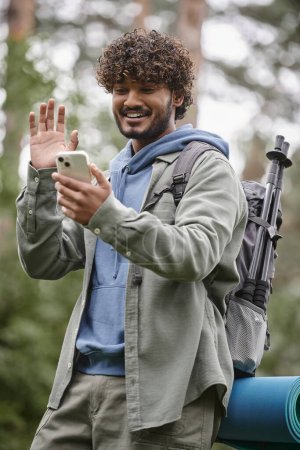Überglücklicher indischer Wanderer mit winkendem Rucksack bei Videoanruf auf Smartphone im Wald