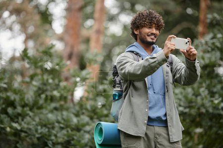 alegre joven senderista indio con mochila tomando fotos en el teléfono inteligente en el bosque borroso