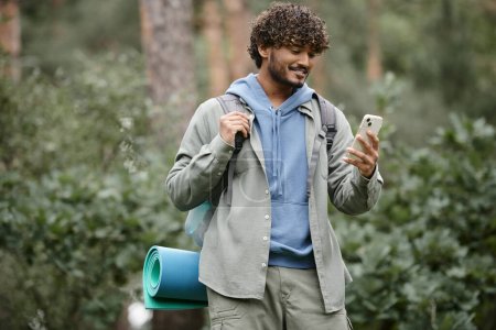 Téléchargez les photos : Jeune touriste indien souriant avec sac à dos en utilisant un smartphone tout en se tenant debout dans la forêt floue - en image libre de droit