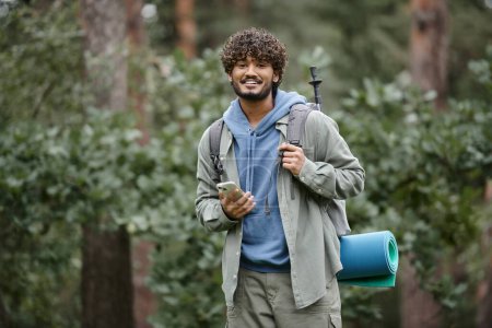 Lächelnder junger indischer Wanderer mit Rucksack, Smartphone in der Hand und Kamera im Wald