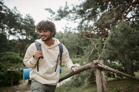 Lächelnder junger indischer Wanderer mit Rucksack läuft in der Nähe von Zaun im verschwommenen Wald