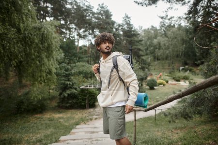 junger indischer Reisender mit Rucksack und Wanderstöcken steht im Wald