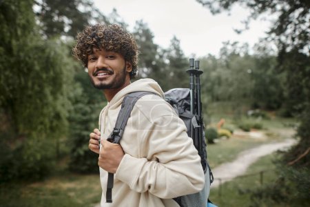 Porträt eines lächelnden jungen indischen Reisenden mit Rucksack, der im verschwommenen Wald in die Kamera blickt