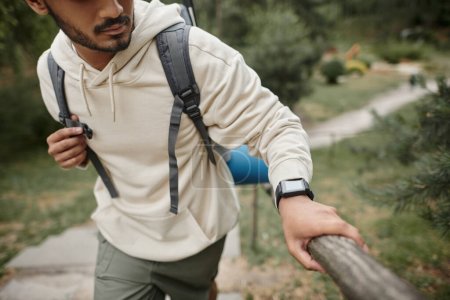 abgeschnittene Ansicht eines jungen indischen Touristen mit Rucksack und Smartwatch, der in der Nähe von Zaun im Wald spaziert