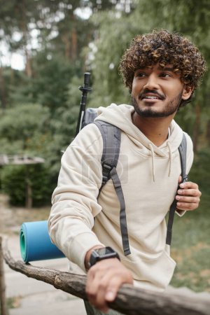 hombre indio positivo con mochila y smartwatch de pie cerca de valla de madera en el bosque