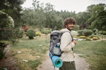 smiling young indian hiker with backpack and trekking poles looking at camera on path in forest