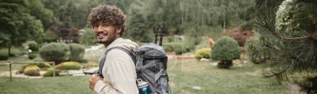 smiling indian hiker with backpack and trekking poles looking at camera on path in forest, banner