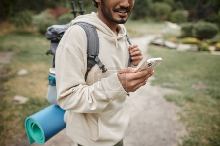 vista recortada de viajero indio sonriente con mochila usando teléfono inteligente en el bosque