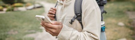 partial view of traveler with backpack using smartphone in blurred park, banner