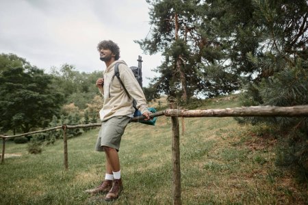 carefree indian hiker with backpack standing near wooden fence in forest