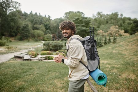 positiver junger indischer Wanderer mit Rucksack blickt in die Kamera mit Landschaft im Hintergrund