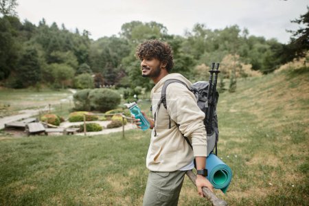 sonriente joven turista indio con mochila sosteniendo botella deportiva cerca de valla al aire libre