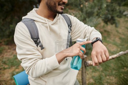 abgeschnittene Ansicht eines lächelnden indischen Reisenden mit Rucksack und Sportflasche mit Smartwatch im Freien