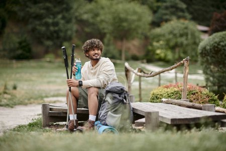 alegre senderista indio sosteniendo bastones de trekking y botella deportiva cerca de la mochila en el bosque