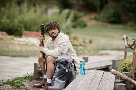 junger indischer Tourist hält Trekkingstöcke in der Nähe des Rucksacks, während er sich im Wald ausruht