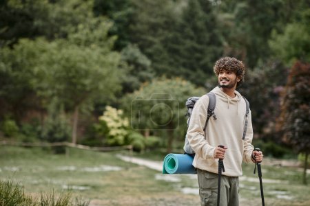 cheerful indian backpacker holding hiking sticks on path in forest, travel and adventure concept