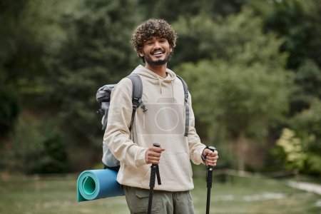 positive indian backpacker holding trekking poles on path in forest, travel and adventure concept