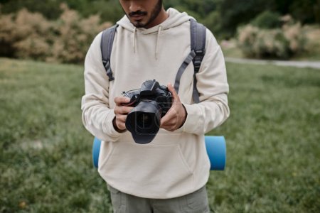 Téléchargez les photos : Vue recadrée du photographe indien en sweat à capuche tenant appareil photo professionnel, sac à dos et voyage - en image libre de droit