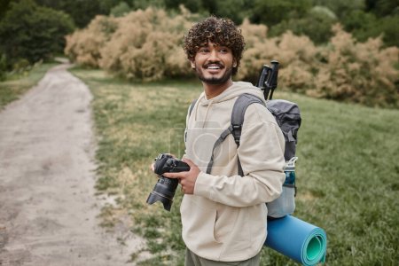 cheerful indian photographer with backpack holding professional camera, adventure and hiking