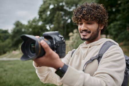 lockige indische Fotografin mit Rucksack, die Fotos mit professioneller Kamera, Abenteuer und Reisen macht