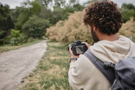 Rückansicht des lockigen indischen Fotografen mit Rucksack, der mit professioneller Kamera im Freien fotografiert