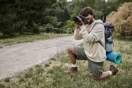 positive indian photographer with backpack taking photo on professional camera, natural location