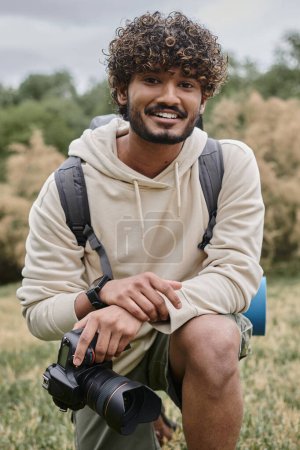 fotógrafo indio alegre sosteniendo cámara profesional y mirando a la cámara en el bosque, vertical