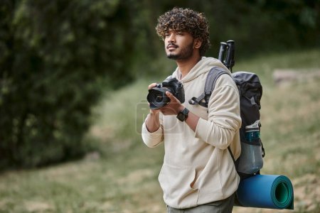 pensive indian tourist holding professional camera, hiker with backpack in forest, explorer