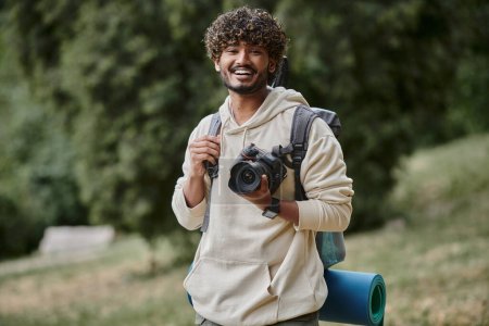 excited indian tourist holding professional camera, hiker with backpack in forest, laughter