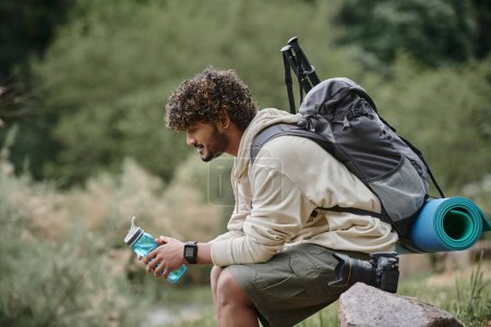 happy indian tourist sitting with backpack and holding bottle with water in forest, adventure