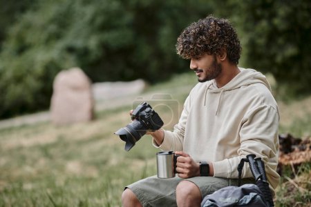 curly indian tourist holding professional camera and sitting on rock, man with thermos cup in forest