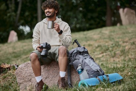 happy indian tourist holding professional camera and sitting on rock, man with thermos cup in forest