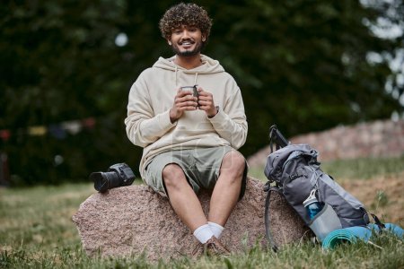 Indianer hält Thermoskanne in der Hand und sitzt auf einem Felsen, fröhlicher Tourist blickt im Wald in die Kamera