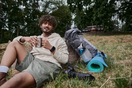indian man holding thermos mug and sitting near camera and backpack with travel gear, tourist