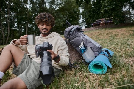 happy indian man holding thermos mug and looking at photos on camera, tourist near travel gear