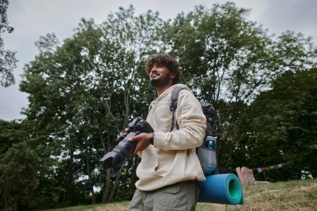 glücklicher indischer Mann mit Digitalkamera und Rucksack im Wald stehend, Fotograf