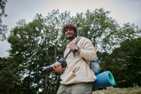 hombre indio alegre sosteniendo la cámara digital y de pie con la mochila en el bosque, fotógrafo