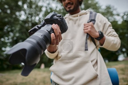 cropped view of indian man holding digital camera and standing with backpack in woods, photographer