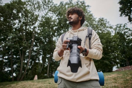 hombre indio concentrado sosteniendo cámara digital y de pie con mochila en el bosque, fotógrafo