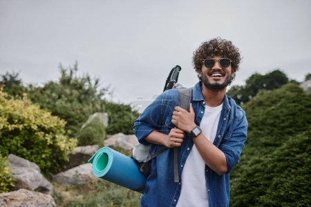 glücklicher indischer Mann mit Sonnenbrille steht mit Rucksack auf grünem Hügel, Wanderer mit Reiseausrüstung