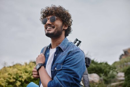 cheerful indian man in sunglasses standing with backpack on green hill, hiker with travel gear