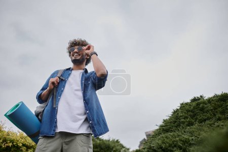 fröhlicher indischer Mann mit Sonnenbrille und Rucksack auf Hügel stehend, Wanderer mit Reiseausrüstung