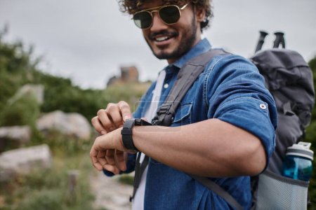 happy indian man in sunglasses and denim shirt checking time on wristwatch, hiker with travel gear