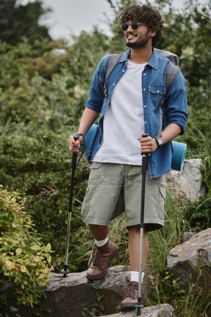 heureux randonneur indien en lunettes de soleil tenant bâtons de randonnée et marche avec sac à dos pendant le trekking
