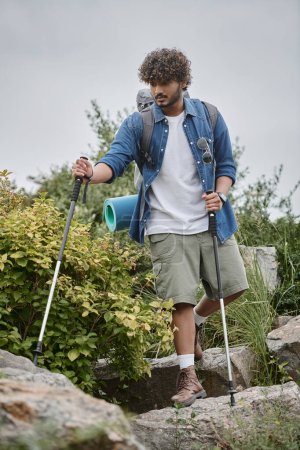 focused indian man walking with backpack and holding hiking sticks during trekking, wild nature