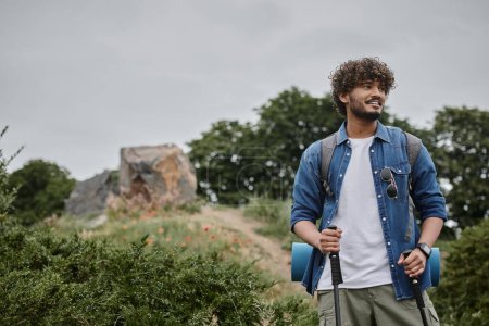 focused indian backpacker holding trekking poles on path in woods, travel and adventure concept